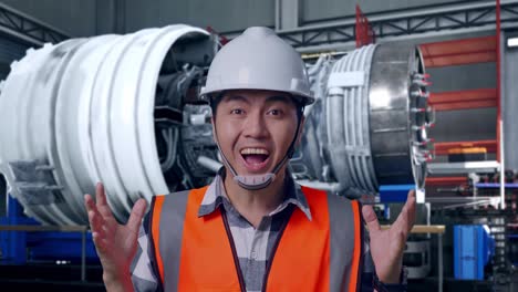 close up of asian male engineer with safety helmet smiling to camera and saying wow while standing with airplane engine maintenance conducted, analytics checking the turbine