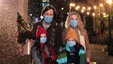 close-up view of caucasian happy family wearing facial masks holding presents and smiling at camera on the street while it¬¥s snowing in christmas