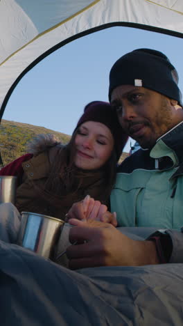 multiethnic backpacker couple sit in tent on mountain hill: they hug and drink tea. two travelers stopped to rest during adventure vacation. romantic hiker family admire the nature. vertical shot