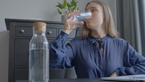 woman indoors drinking a giant glass of water with a laptop on the table