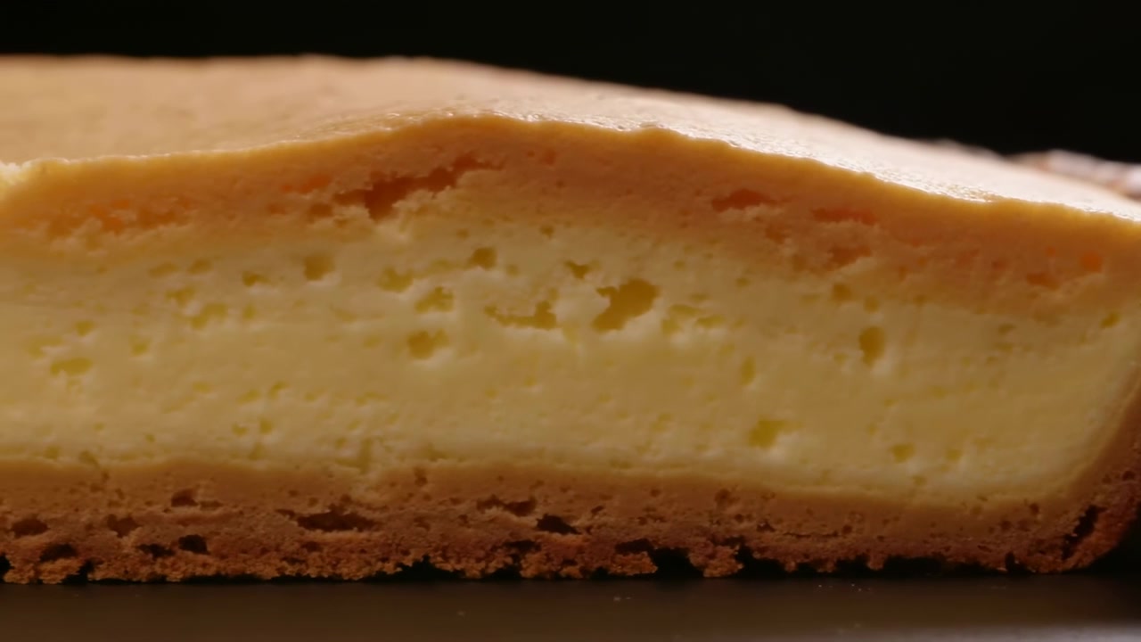 Ripresa in movimento quasi a livello che rivela una spessa fetta di cheesecake cotta in sezione trasversale, con superficie dorata, interno cremoso e poroso, piccole bolle d'aria, crosta croccante e dorata, e illuminazione da studio soffusa.