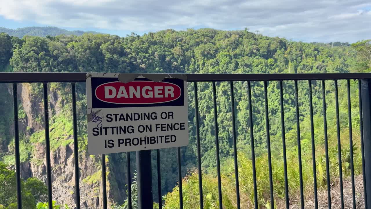 A stationary camera captures a metal fence with a danger sign warning against standing or sitting, overlooking a sunlit forested cliffside under clear daylight