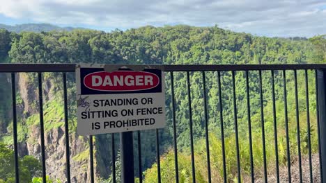 a stationary camera captures a metal fence with a danger sign warning against standing or sitting, overlooking a sunlit forested cliffside under clear daylight