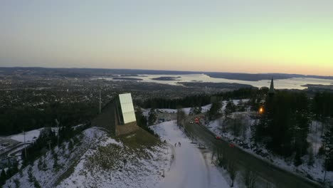 Oslo-cityscape-drone-dolly-shot-with-Holmenkollen-ski-jump-in-foreground,-Holmenkollbakken,-Vinterpark-Winterpark-Tryvann-Past-Ski-Jump-at-Sunset