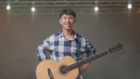 adolescente asiático con guitarra poniendo las manos en la cadera y sonriendo en el escenario