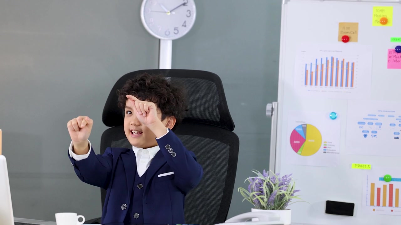 A young boy in a blue suit gestures confidently while sitting at an office desk