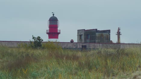 Faro-De-Puerto-Pintado-De-Rojo-En-El-Puerto-De-Liepaja-En-Un-Día-Nublado,-Vista-Lejana,-Tiro-Medio-De-ángulo-Bajo-Sobre-Las-Dunas-Costeras-Con-Arena-Blanca