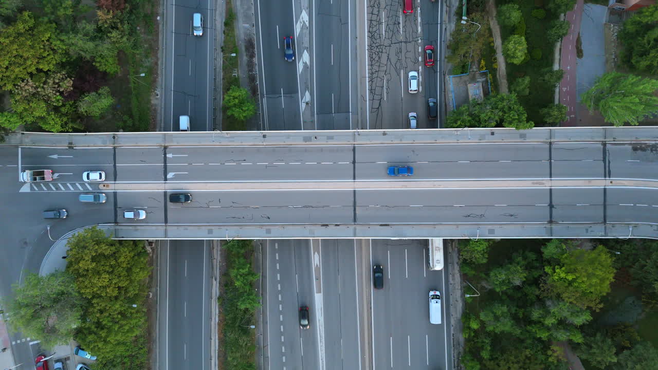 Vertical drone shot capturing fluid traffic on a busy highway interchange and overpass. Vehicles smoothly passing on multiple lanes, surrounded by lush greenery, emphasizing urban transportation