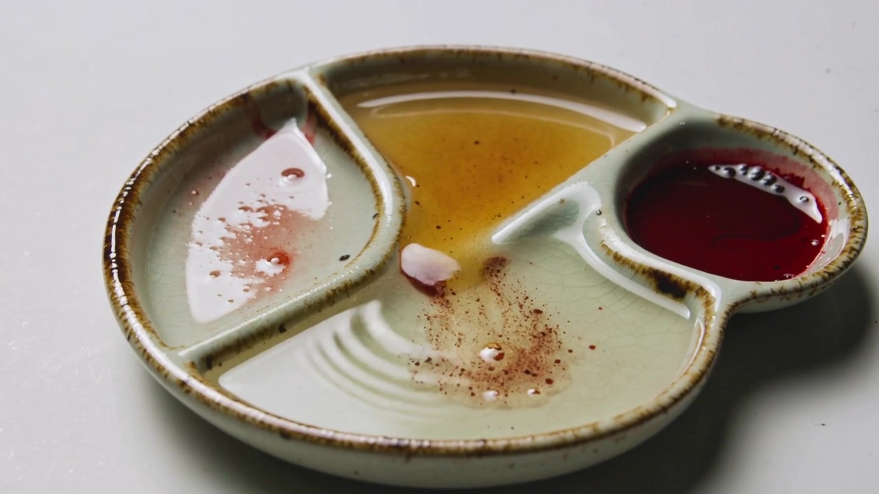 Líquidos ondulando em uma paleta de cerâmica dividida sobre a mesa após a queda de uma pétala, com âmbar e vermelho se misturando.
