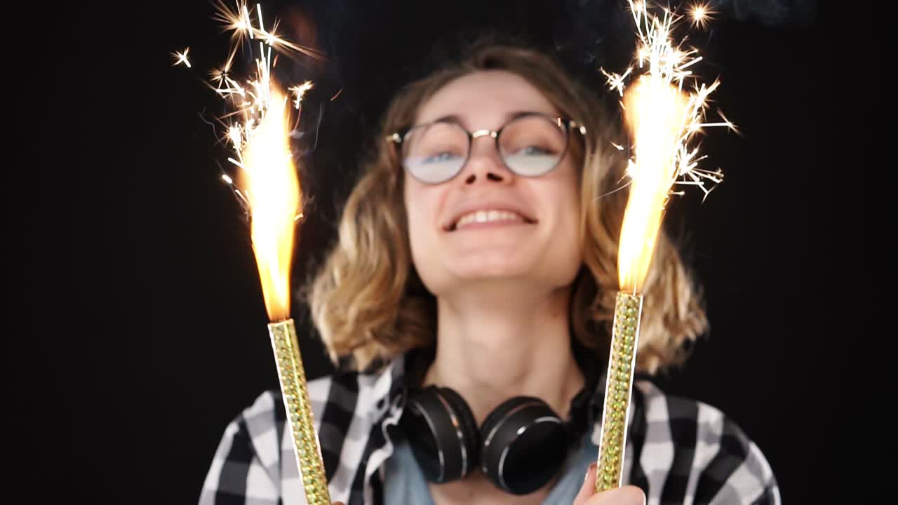 Close up european hipster girl in black and white plaid shirt, headphones on neck celebrating with sparklers and bengal fire. Happy woman smiling girl holds two sparklers close to the camera against black background. Slow motion