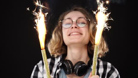 chica hipster europea en camisa a cuadros blanca y negra, auriculares en el cuello celebrando con chispas y fuego de bengala. mujer feliz sonriendo chica sostiene dos chispas cerca de la cámara contra un fondo negro. cámara lenta