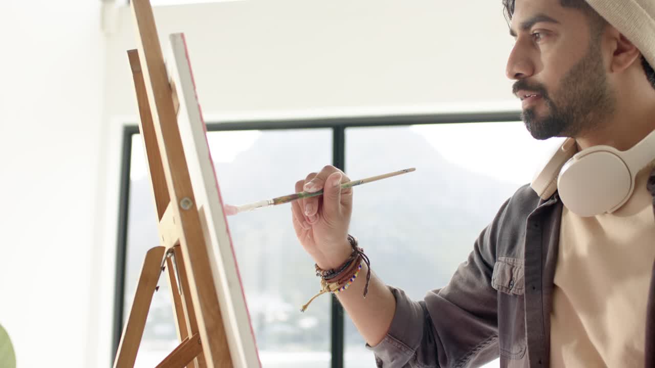 Indisk mandlig kunstner står foran et tomt lærred i studiet og løfter penslen mod staffeliet for at påføre strøg