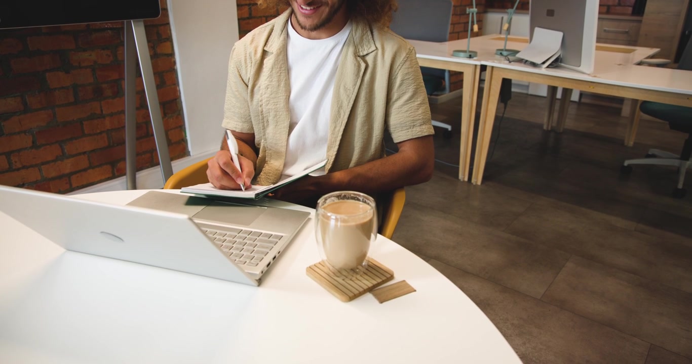 Headphones playing lecture, adult man writing in notebook using open laptop and mug in coworking