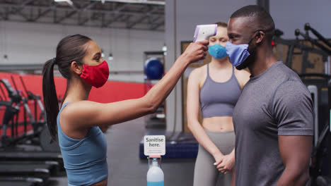 Mujer-Caucásica-En-Forma-Con-Máscara-Facial-Midiendo-La-Temperatura-De-Un-Hombre-Afroamericano-En-Forma-En-El-Gimnasio