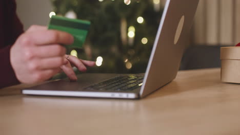 camera focuses on man's hands using laptop and holding credit card sitting at a table near a present in a room decorated with a christmas tree