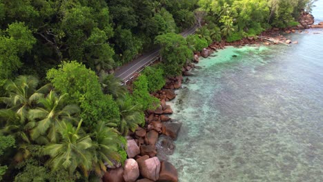 Bird-eye-drone-shot-of-passing-bus-on-asphalt-road,-granite-rocks,-trees-and-turquoise-water-on-the-baie-lazare-shore,-Mahe-Seychelles-30-fps
