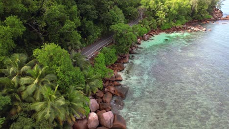 bird eye drone shot of passing bus on asphalt road, granite rocks, trees and turquoise water on the baie lazare shore, mahe seychelles 30 fps