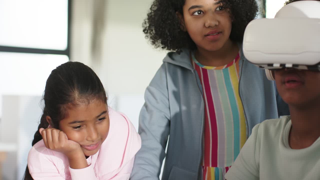 African American girls at table left in pink curiosity urging right girl using white VR headset
