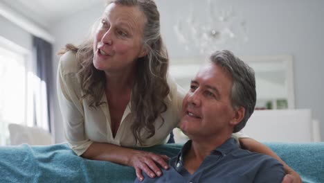 happy senior caucasian couple watching tv in living room and talking, wife embracing sitting husband