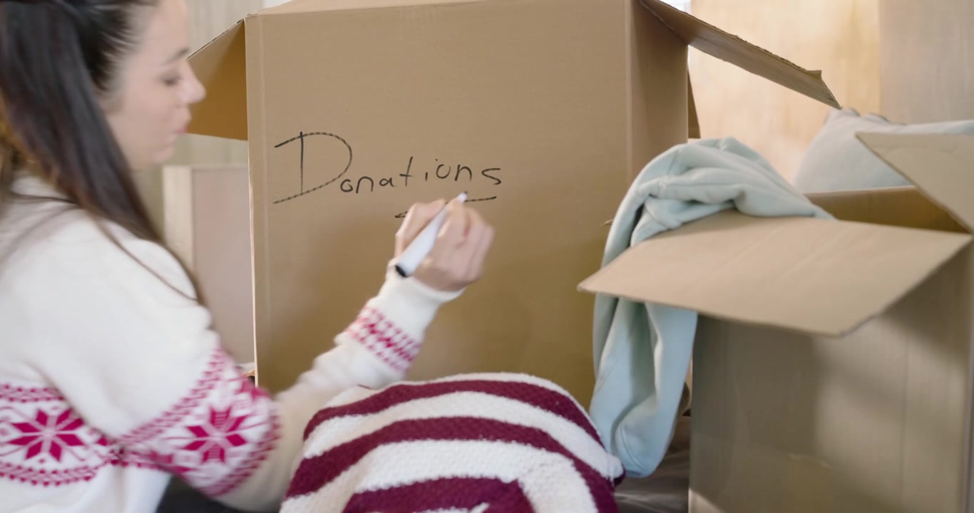 Woman in mid-20s writing Donations label with black marker, picking sweaters and packing box on bed