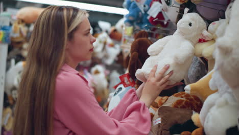 lady in pink dress inspects a white plush toy in a toy store, focused and examining it, surrounding her are other colorful stuffed toys neatly arranged on shelves in a brightly lit store