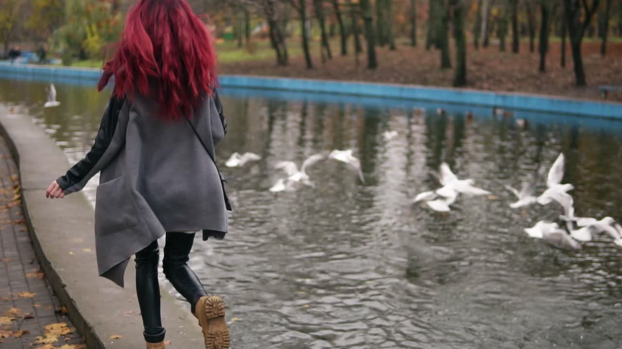Back view of young woman with red hair running towards gulls in an artificial pond and making them fly away. Autumn day in park