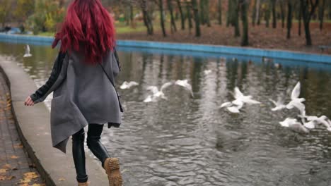 Back-view-of-young-woman-with-red-hair-running-towards-gulls-in-an-artificial-pond-and-making-them-fly-away.-Autumn-day-in-park