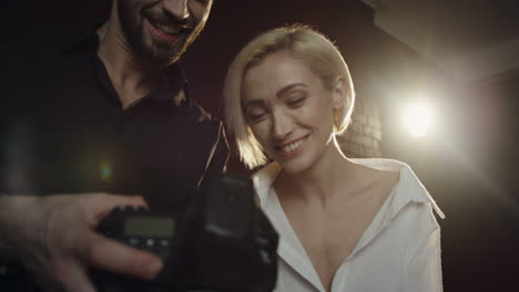 bottom view of a photographer man showing photos of photoshoot to the young blond woman model on the little camera screen in the dark studio