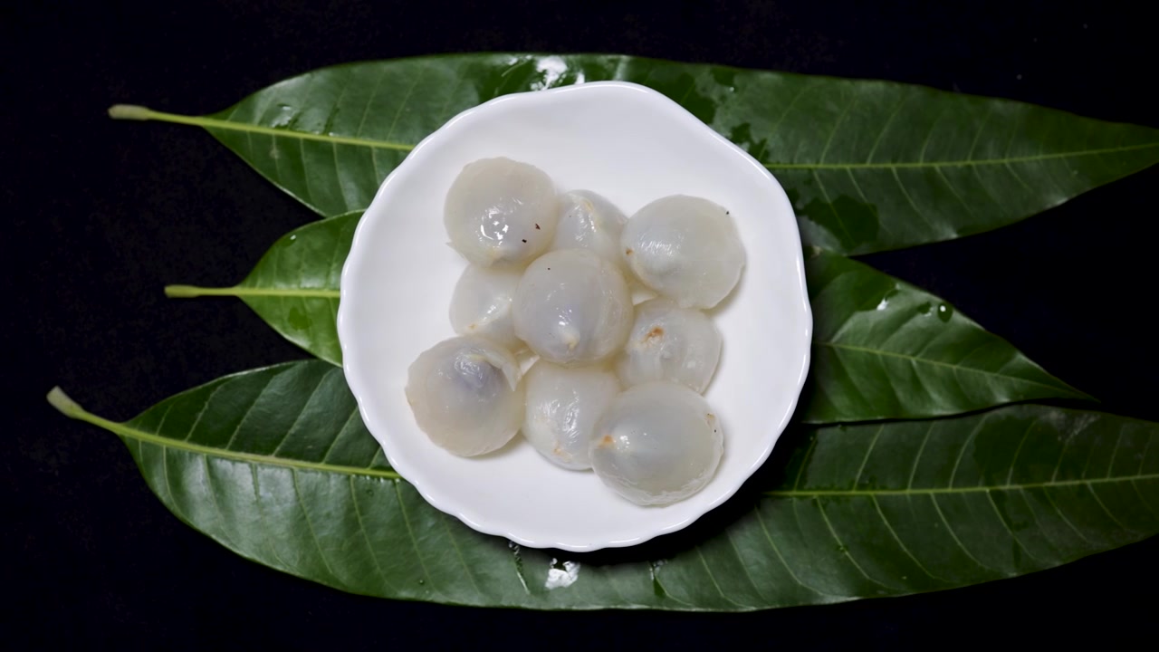 Cùi vải mọng nước được phục vụ trong bát sứ với nền là tán lá nhiệt đới.