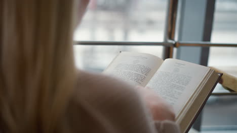 close-up rear view of lady flipping to new page in book with tea cup in front, blurry busy urban settlement visible in background through the window