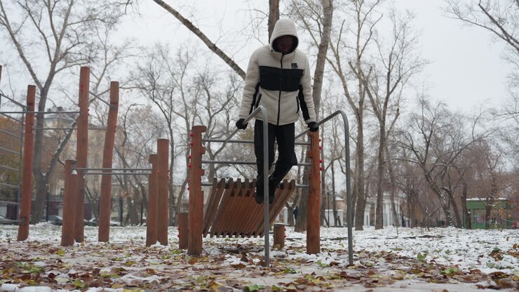 Young adult performs intense muscle-building workout on parallel bars in snowy urban park, dressed in a warm hoodie and gloves, he lifts himself in a dip exercise as people walk in the distance