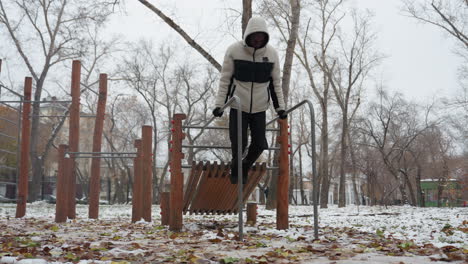 young adult performs intense muscle-building workout on parallel bars in snowy urban park, dressed in a warm hoodie and gloves, he lifts himself in a dip exercise as people walk in the distance