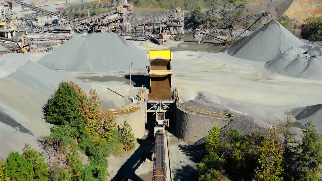 Conveyor belts and gravel piles surround dump truck actively pouring fill into hopper for processing along conveyor belt, aerial establish