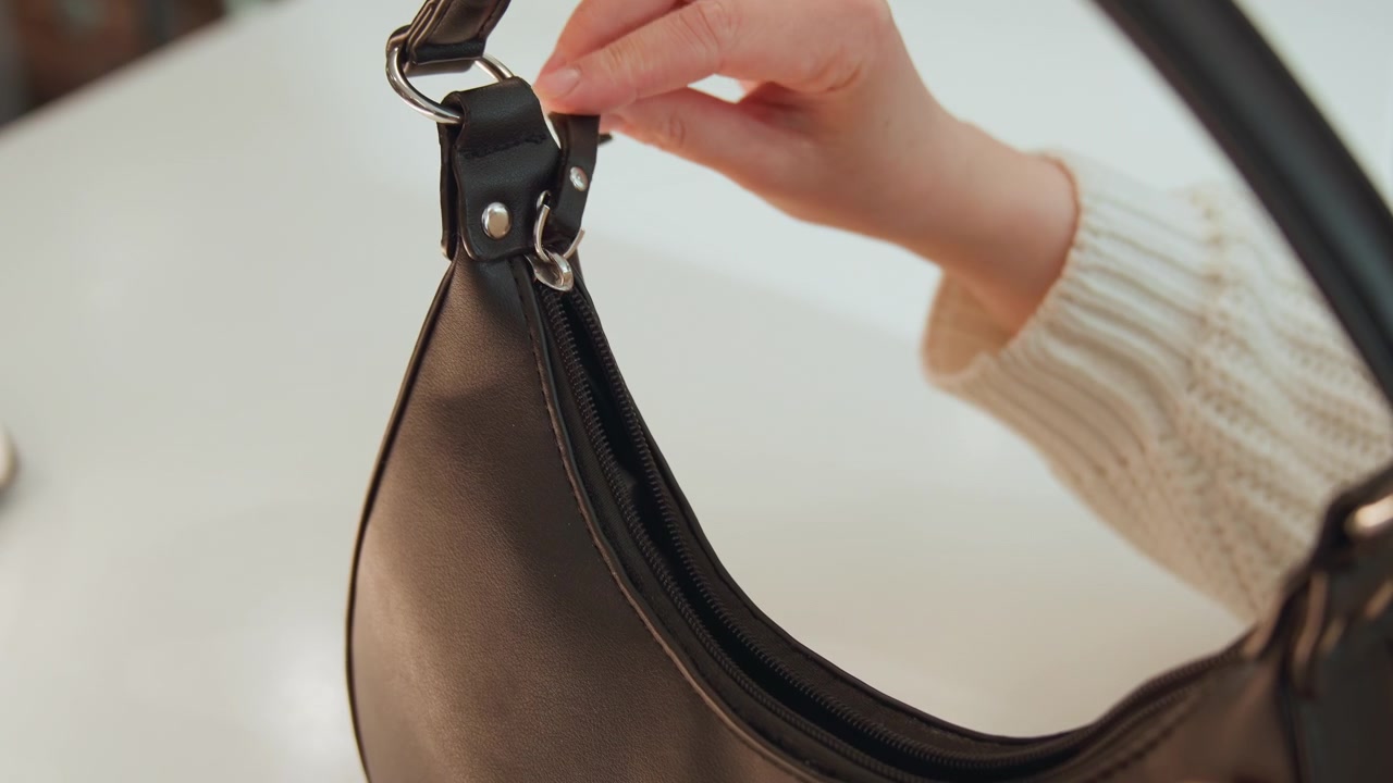 Primo piano delle mani che maneggiano una borsa in pelle nera, con le dita che chiudono la fibbia metallica, regolano la tracolla, sollevano il bordo per rivelare il tiretto della cerniera e dondolano delicatamente la borsa durante un'attenta ispezione in una luce soffusa