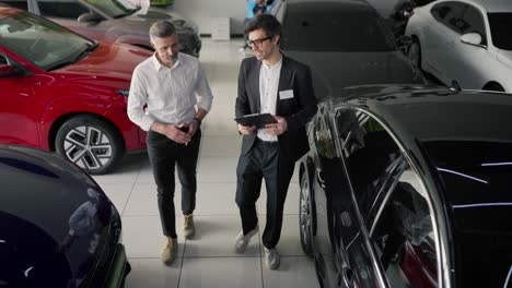 Top-view-of-a-confident-brunette-male-assistant-in-a-business-suit-talking-to-a-middle-aged-man-in-a-white-shirt-about-modern-car-models-and-talking-about-them-in-a-car-showroom