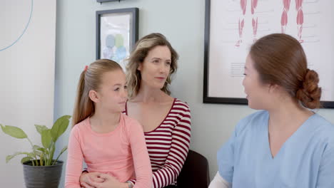 happy diverse girl patient sitting with mother and female nurse talking in hospital, slow motion