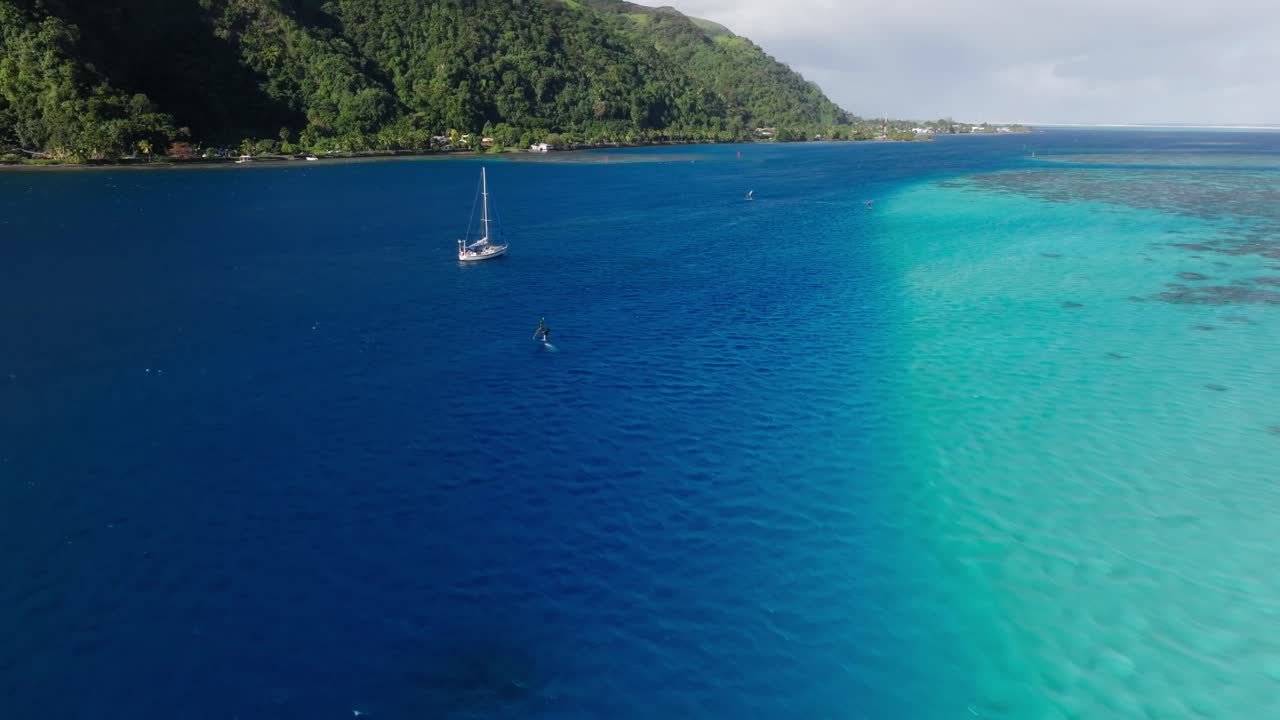 Following a windsurfer while panning up the drone camera to show the mountainous coast. Captured in Tahiti, French Polynesia