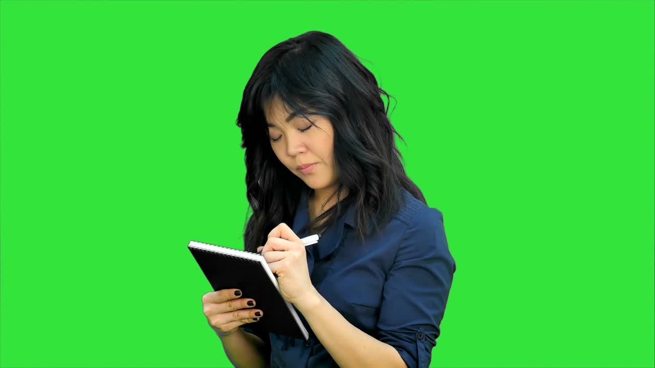 Tankefull asiatisk kvinne tar notater i en notatblokk på en grønn skjerm, Chroma Key.