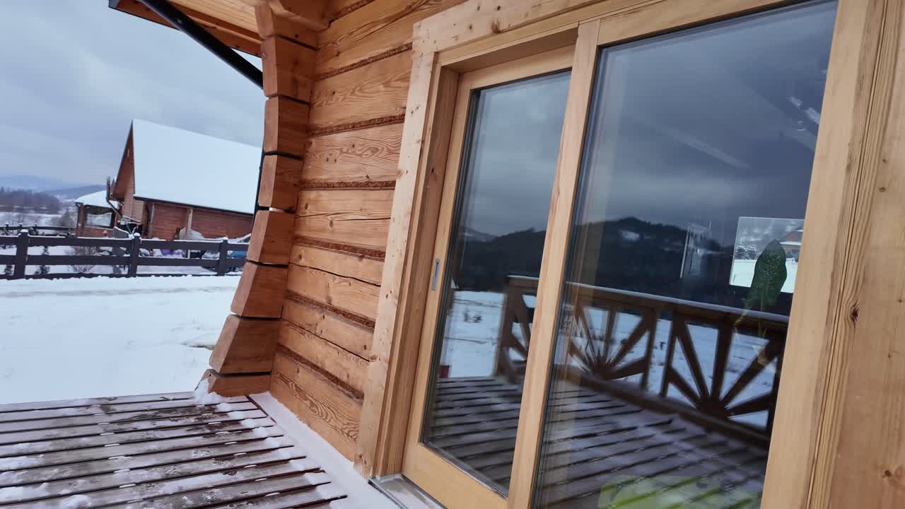Vista invernale di una terrazza in legno che termina con una porta a vetri dove le montagne innevate appaiono riflesse