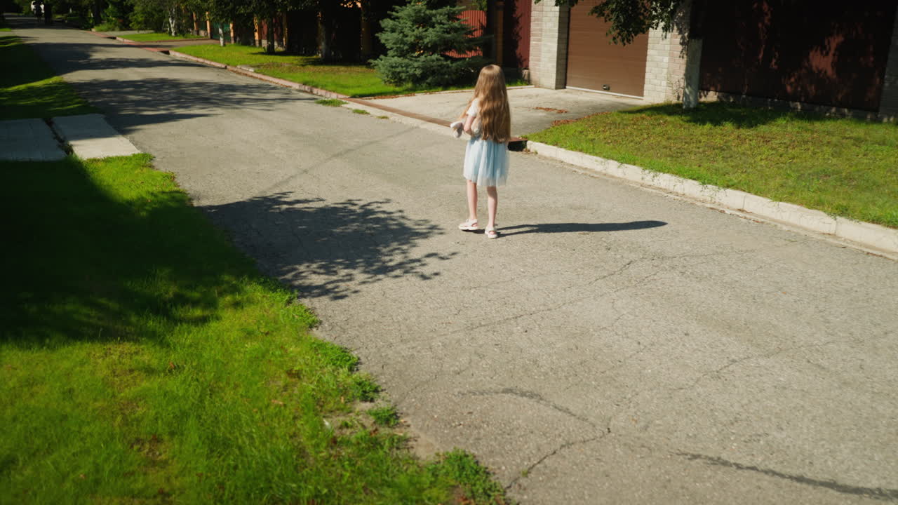 Girl in gown walking alone along cracked suburban road under bright sun, holding stuffed toy, surrounded by green grass, sidewalks, driveways, and residential fences in peaceful neighborhood