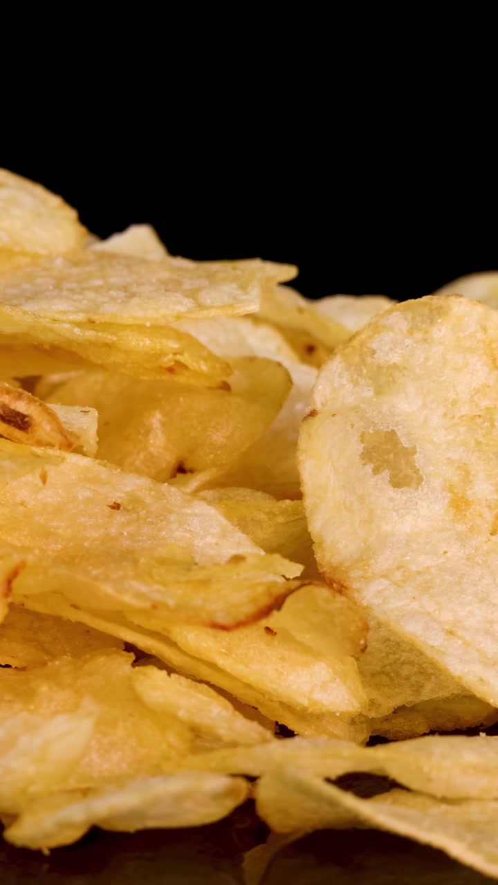 Camera zooms out from close-up of crispy potato chips, dramatic lighting, black background, reflective surface