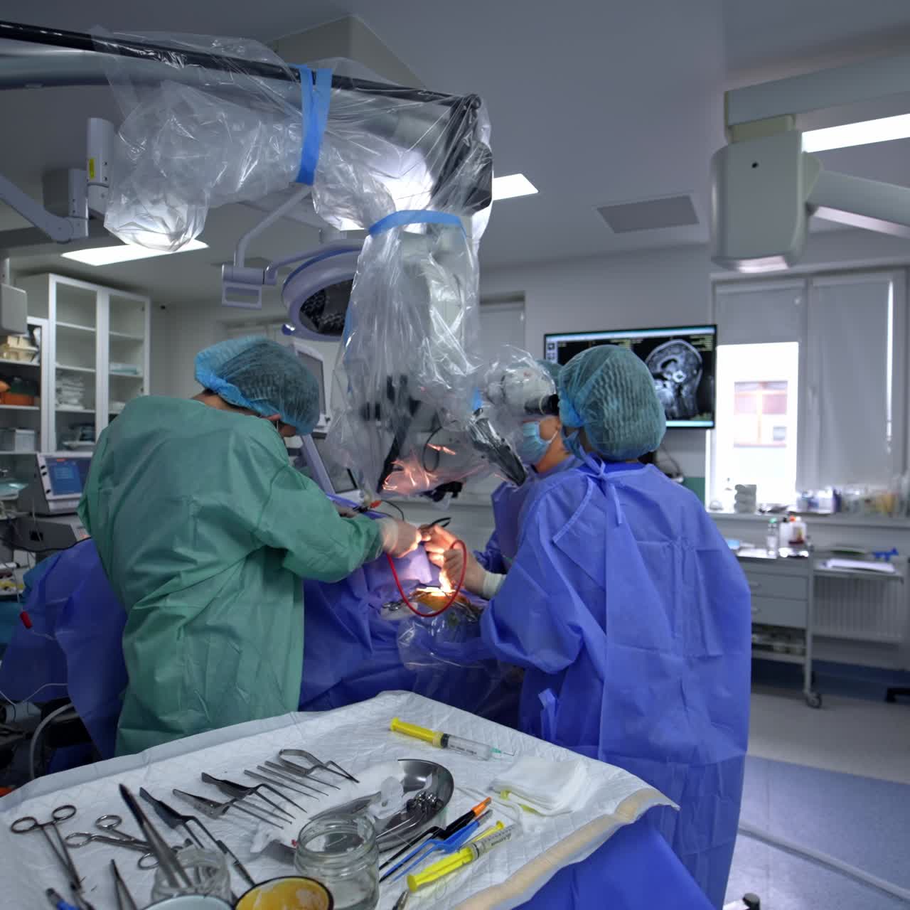 Male assistant passing the instruments to the main doctor taking them from a table. Neurosurgeon sits at backdrop looking at microscope