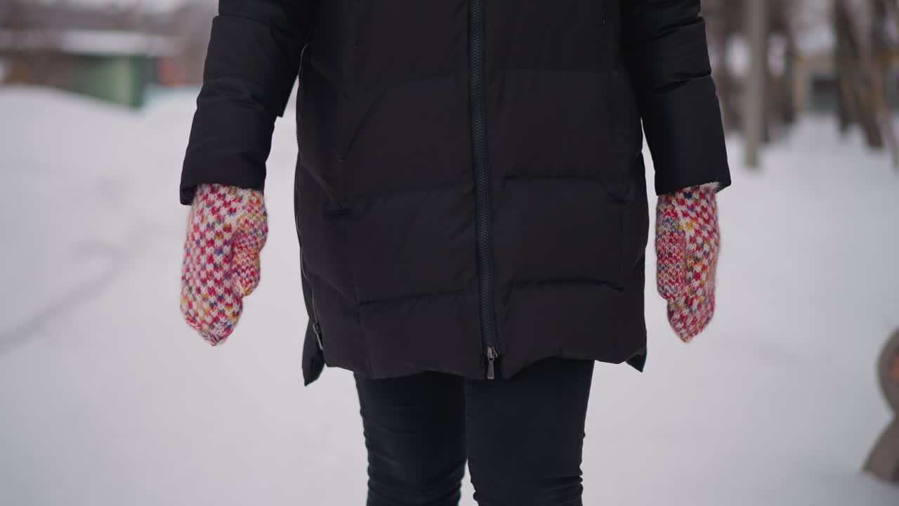 Female wearing black winter coat walking outdoors in snowy environment with both hands visible in colorful knitted mittens close up detail lower body cold frosty season lifestyle clothing urban background