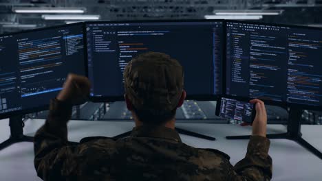 back view of happy asian military looking at code on mobile phone while write code with multiple computer screens in data center