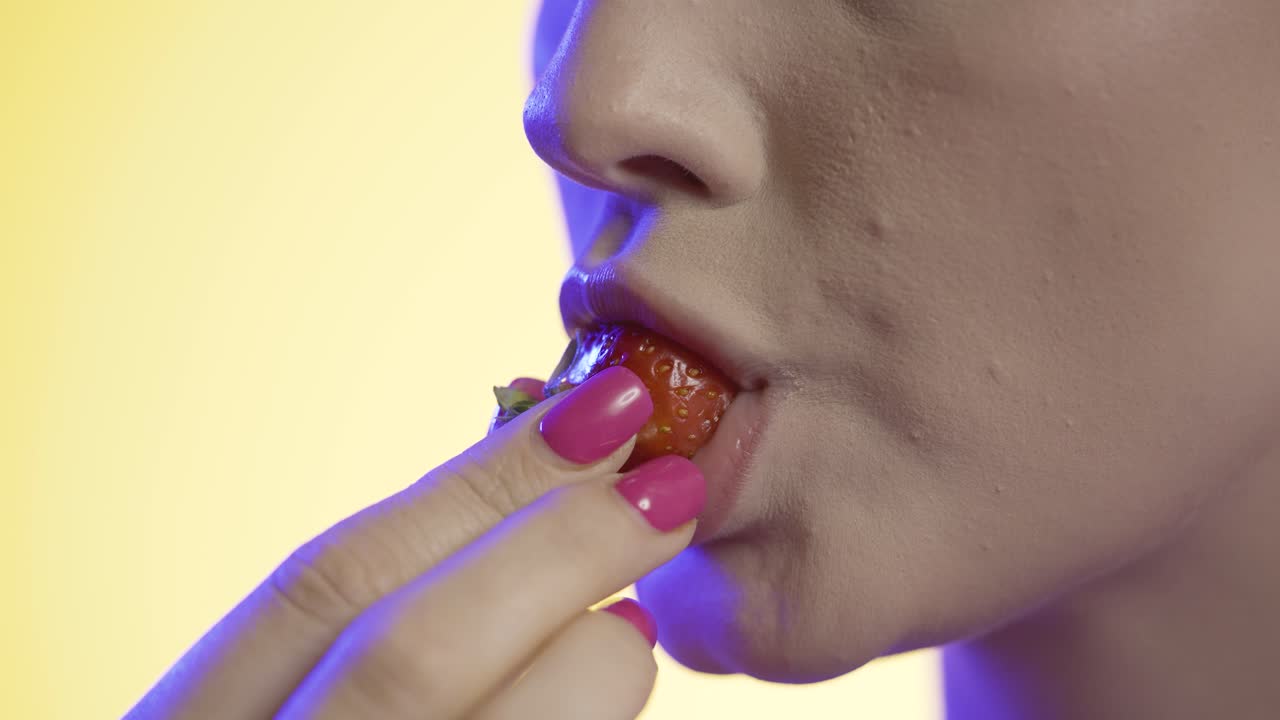 молодая женщина ест клубнику, жует рот крупным планом на желтом фоне, розовое освещение
