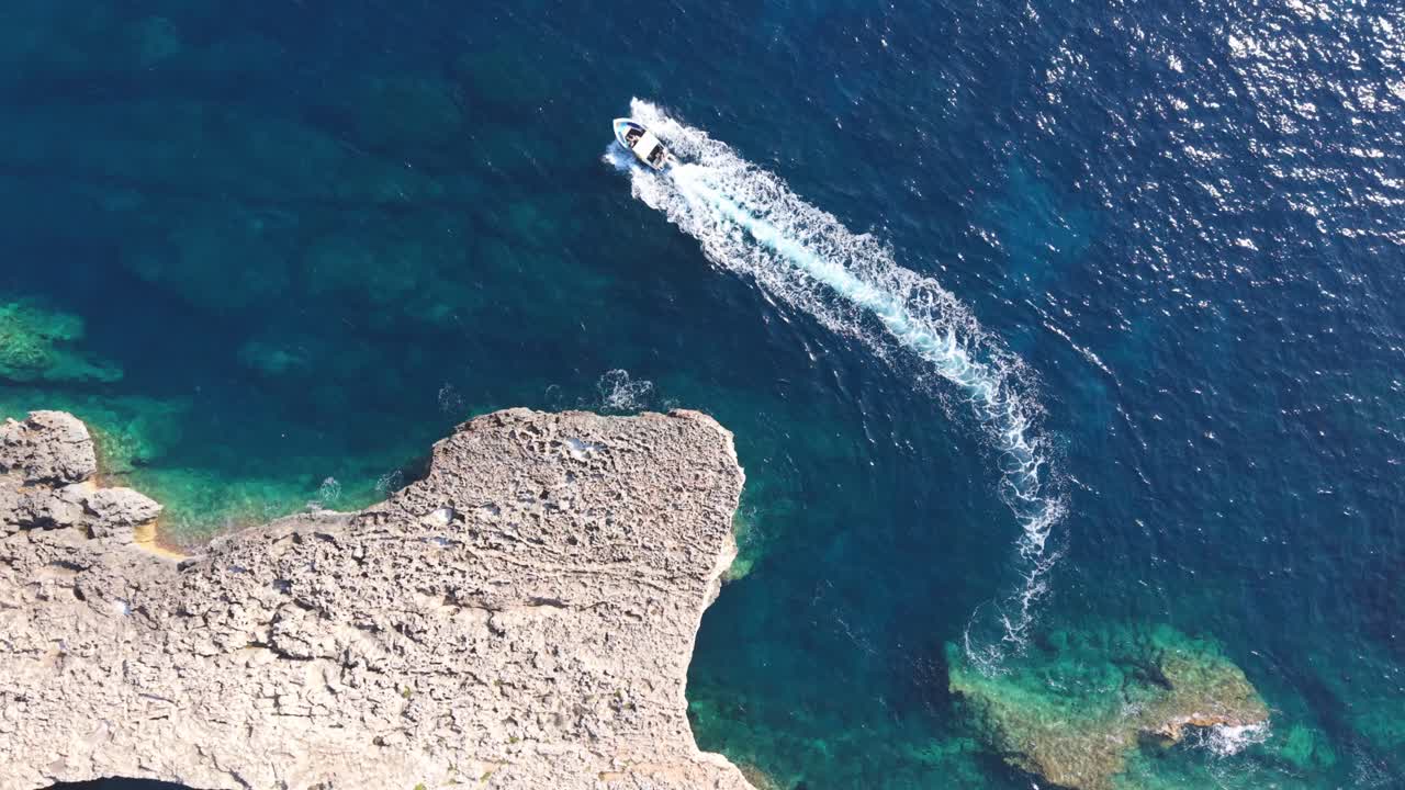 A drone perspective shows a small speedboat leaving a white trail on the deep blue sea near a rugged rocky cliff. The crystal-clear turquoise waters below reveal underwater rocks and coastal textures