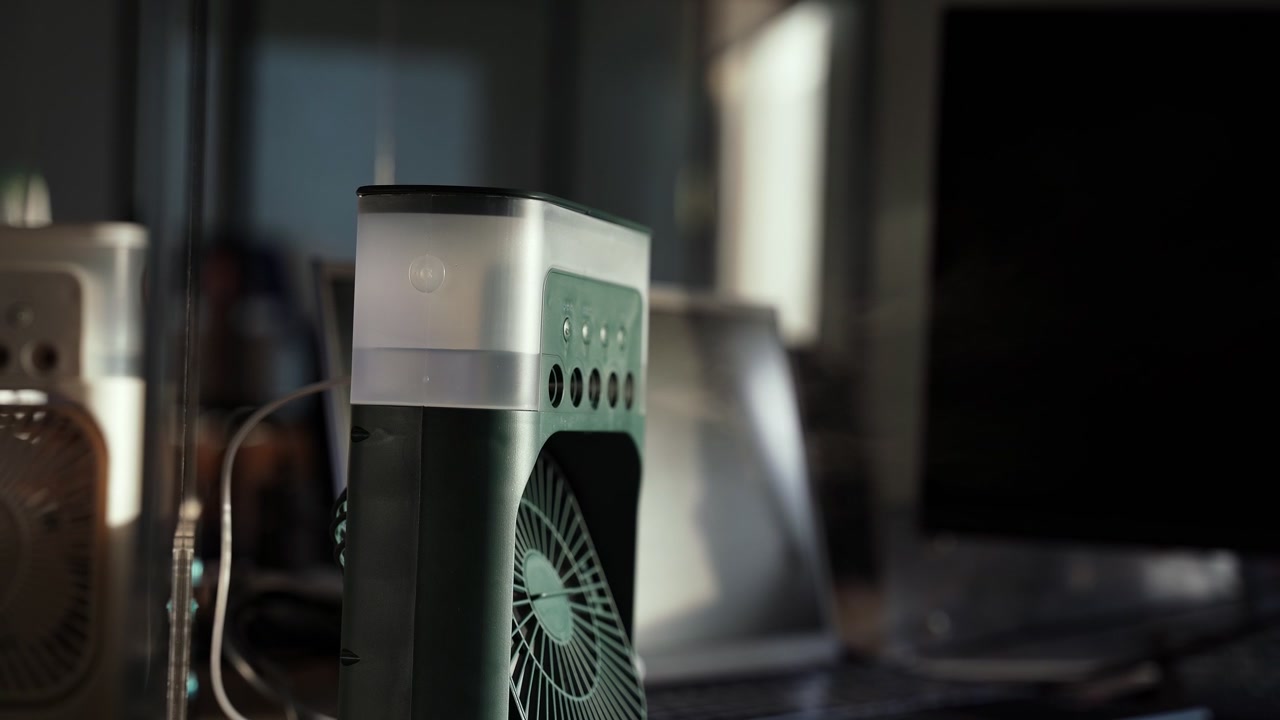 Primo piano di un ventilatore raffrescatore d'aria portatile da tavolo con serbatoio d'acqua, posizionato su una scrivania scura accanto a un laptop e un monitor, in un'illuminazione interna suggestiva
