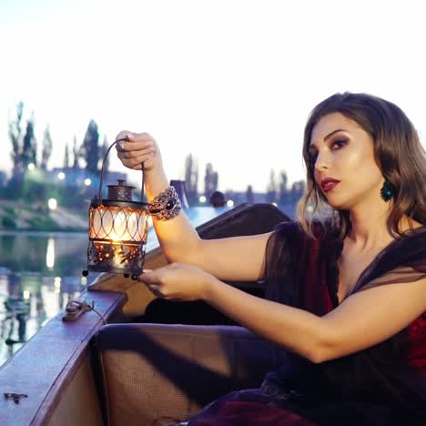 a beautiful model in black and red dress is holding a decorative lantern in her hands and posing on the gondola in the evening on the background of water and sky. carnival in venice. blurred background