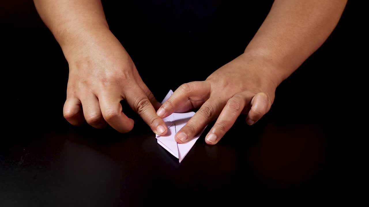 Adult hands carefully fold white paper into origami bird, overhead view, dramatic lighting