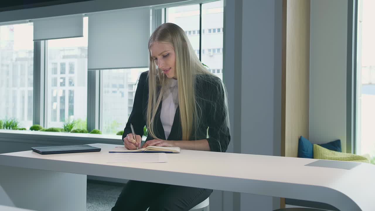 Donna elegante che lavora in un ufficio di stile. Una moderna donna bionda in un tailleur alla moda è seduta a un tavolo in un luminoso ufficio contemporaneo e scrive su dei documenti.